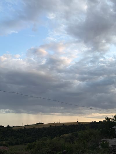 The image shows a view of rolling hills covered with trees and fields under a dramatic cloudy sky. In the foreground, there is dense green vegetation and what appears to be part of a building on the right. A dark wire runs horizontally across the middle of the frame, bisecting the sky and the landscape. In the distance, atop the hills, there are a few scattered structures, possibly including utility poles and a solitary tree. There is a visible rain shower in the far distance, indicated by streaks of precipitation against the lighter sky. The overall scene suggests an overcast or approaching storm over a rural or semi-rural area. This image was taken in the city of Nikopol, Bulgaria.