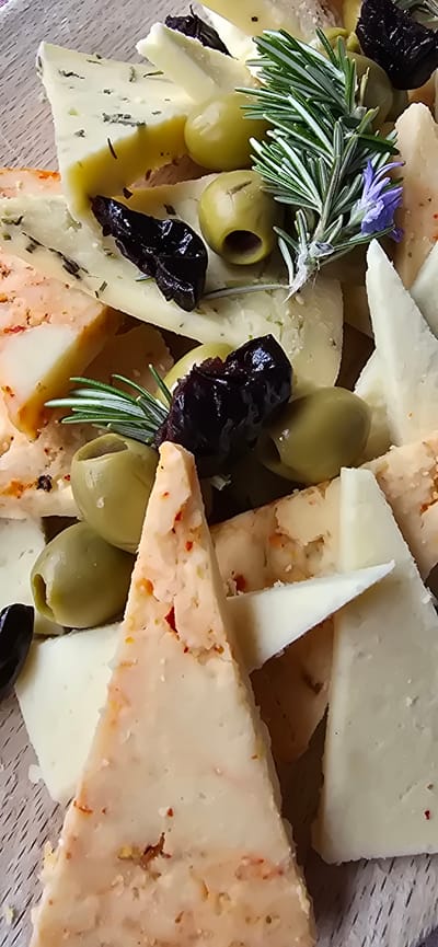 The image depicts a cheese board featuring a variety of cheeses cut into triangular shapes. The cheeses vary in texture and color, some appearing plain white, while others have herbs or chili flakes incorporated. Green and black olives are scattered among the cheese pieces. A sprig of rosemary with a small purple flower is also present. The food appears to be arranged on a wooden board. The location in the city of Kopito, Montenegro is not discernible from the image. There is no information about people, actions, or interactions depicted in the image.