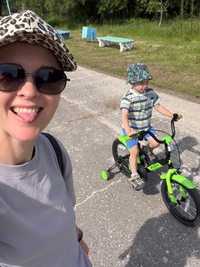 In Kul'dur, Russia, a woman with a leopard-print hat and sunglasses sticks her tongue out at the camera, her mouth wide open in a smile. She wears a gray t-shirt and a black backpack strap is visible over her shoulder. To her right, a young boy wearing a green bucket hat and a striped t-shirt rides a bright green bicycle with training wheels on an asphalt path. The boy is wearing blue shorts and sneakers. The background features a grassy area with trees and some outdoor furniture, including a light blue structure and a bench. The lighting suggests it is a sunny day.