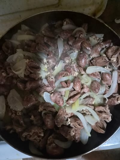A close-up, overhead shot shows chicken hearts and sliced onions being stir-fried in a black, non-stick frying pan. The pan is on a stovetop, with a portion of the beige stovetop visible at the bottom of the frame. The hearts are dark red and irregularly shaped, while the onions are translucent white and sliced into crescent shapes. Steam rises from the pan, indicating the food is hot and being actively cooked. The lighting is dim, likely from an overhead kitchen light. The image appears to have been taken in a domestic kitchen in Kaleytsa, Bulgaria.