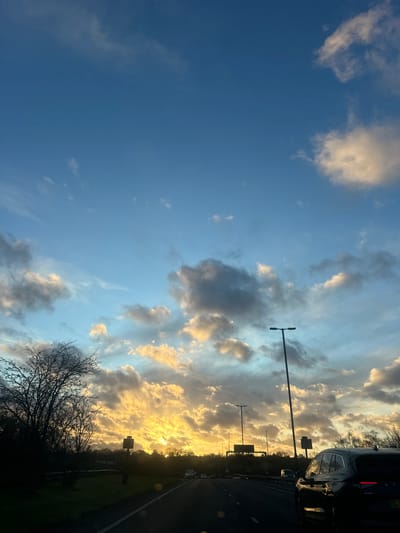 The scene is observed in Upper Upnor, United Kingdom, during the golden hour, likely sunset, under a partly cloudy sky. The upper portion of the sky is clear blue, transitioning to a mix of scattered cumulus and stratocumulus clouds below. These clouds are intensely illuminated with golden-orange light from the low sun on the horizon, while other cloud formations appear darker. A multi-lane paved road extends into the distance, marked by a solid white line on the immediate left lane's edge. In the right foreground, the rear of a dark-colored Skoda Enyaq SUV is partially visible. Multiple other vehicles appear as dark silhouettes further down the road, indicating traffic flow. Several tall, dark streetlights/lampposts and a gantry structure with potential road signs are positioned along the roadside. A roadside camera or sensor unit is mounted on a pole on the left. Deciduous trees with bare branches are prominently silhouetted against the bright horizon on the left and along the distant tree line. No individuals are distinctly visible in the frame.