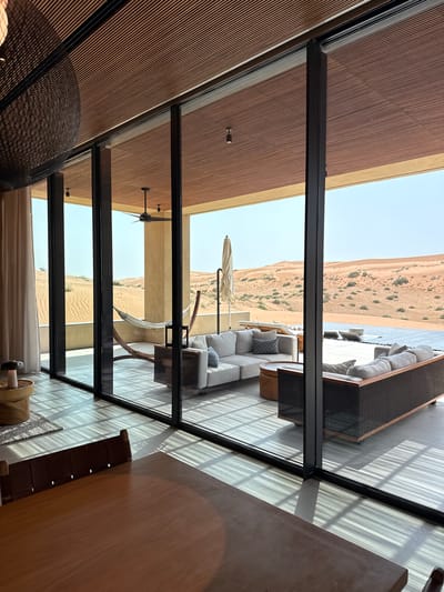 The image shows a modern living area with large glass doors opening onto an outdoor patio and a desert landscape in Ras Al-Khaimah, United Arab Emirates. Inside, a wooden dining table with a dark leather-backed chair is in the foreground. The living area features two white sofas, one with woven sides, and several patterned throw pillows. A round, leather-topped ottoman sits between the sofas. A hammock is suspended on the patio, along with a ceiling fan and an umbrella. The exterior view reveals rolling sand dunes, sparse vegetation, and a swimming pool. Sunlight casts striped shadows across the tiled floor.