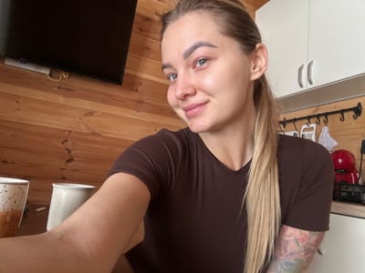 The scene is set indoors, within a location specified as Доња Брезна, Montenegro.
**Subject:**
A female individual is centrally positioned, facing the camera with a subtle smile. She has light skin, light-colored eyes, and light brown or blonde hair styled in a ponytail. Her eyebrows exhibit evidence of permanent cosmetic enhancement. She is wearing a dark brown, short-sleeved top with a V-neck. A floral tattoo is visible on her left forearm/bicep, partially extending from her sleeve. Her right arm and elbow are partially visible on the right side of the image.
**Foreground/Midground Objects:**
On a dark surface in the lower left, two ceramic mugs are present: one with a speckled white and orange/brown pattern, and another plain off-white. Between these mugs, a small, white cylindrical object rests.
**Background Environment:**
The walls in the background are constructed of horizontal light brown wooden planks.
To the left, a large, black flat-screen television is mounted on the wooden wall. Below the television, a white power strip with a blue indicator light and a coiled yellow cable are visible against the wooden surface.
To the right, white upper kitchen cabinets with two silver, curved handles are visible. Below these cabinets, a black metal rod with multiple hooks is mounted on the wooden wall. Hanging from these hooks are various kitchen items, including a white grater and a white patterned cloth. On a light-colored countertop beneath the hooks, a red appliance, consistent with a single-serve coffee machine, is present next to a smaller black appliance with partially legible text that reads "AMY".
**Actions/Interactions:**
The female subject is actively engaged in a self-portrait, looking directly at the camera.