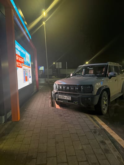 The image captures a scene in Bolshiye Dvory, Russia, at night. A light grey car, identified as a "TOUR" model by the lettering on its front grille, is parked on the side of a brick-paved road. The vehicle's license plate reads "A030PX 797 RUS," indicating its Russian registration. Next to the road, an illuminated advertising board displays text in Russian. Several streetlights are visible in the background, adding to the overall brightness of the scene.