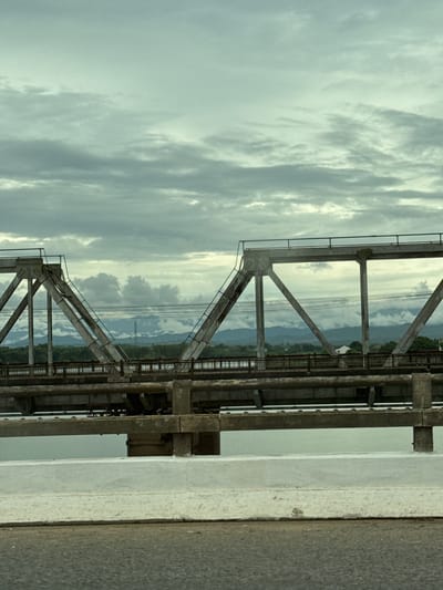 An overpass spans a body of water under a cloudy sky. The overpass is a truss bridge made of concrete and metal. The concrete pylons supporting the bridge are massive, and the metal beams are angled to form triangular supports. A lower deck of the bridge is visible beneath the main span, with a railing along its edge. Trees and distant mountains can be seen on the horizon beyond the bridge. The water beneath the bridge is calm and reflects the overcast sky. The foreground shows a section of a road, likely leading onto the overpass, with a concrete barrier separating it from the water. The overall impression is one of a functional, perhaps aging, piece of infrastructure in a natural setting. This scene is located in Quảng Trị Ward, Vietnam.