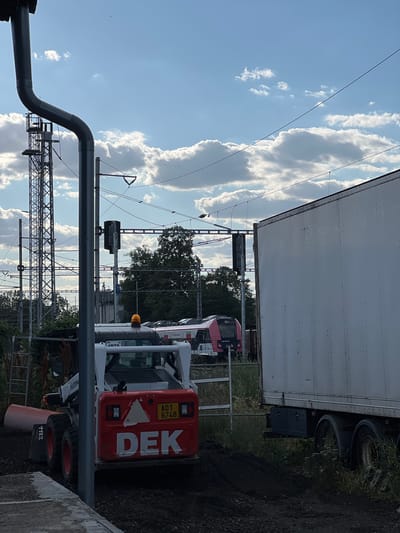 The image shows a construction site in Újezd u Brna, Czechia. In the foreground, a small orange and white skid steer loader with the text "DEK" on its rear is parked on dark soil. The loader has its engine running, indicated by a flashing orange light on top. Behind the loader, a white semi-trailer truck is parked. In the background, a red and white train is visible on a track, alongside overhead power lines and a lattice tower. The sky is blue with scattered white clouds, and there are trees on the horizon. The overall scene suggests ongoing construction or maintenance work near a railway line.