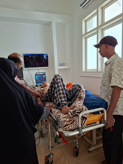 Location: Uleereuleung, Indonesia. An elderly person is undergoing an ultrasound examination in a medical room. A second person is assisting the patient and a third is performing the examination. Two ultrasound monitors are visible on the wall. A walker with wheels is near the patient. The room is tiled with white walls. A skylight is present.