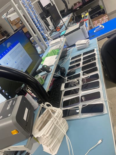 The image displays an indoor industrial or technical workspace located in Amadora, Portugal, featuring multiple workstations with light blue anti-static surfaces. In the foreground, a workstation is equipped with a desktop computer, its monitor displaying a Windows operating system and an open application window. To the left, a grey Zebra label printer exhibits a label with the date "10.11.2023.01." A black flexible arm, a partially visible green Super Bock can, and a can of "TOP GUN" spray are also present near the monitor. The desk surface further includes a black keyboard, a mouse, headphones, and a barcode scanner. To the right, twenty smartphones, primarily black, are arranged face-up in four rows of five, resting on protective bubble wrap. Multiple white charging cables connect to these phones, with additional cables coiled on the desk. Stacks of white paper or cardboard inserts and wrapped white rectangular items, likely electronic components, are visible, along with a green microfiber cloth. In the background, additional workstations are discernible, featuring computer monitors, a silver laser engraving machine with blue coiled tubing, and a blue conveyor belt. Various boxes and equipment are also present in the rear of the facility. No individuals are visible in the scene.