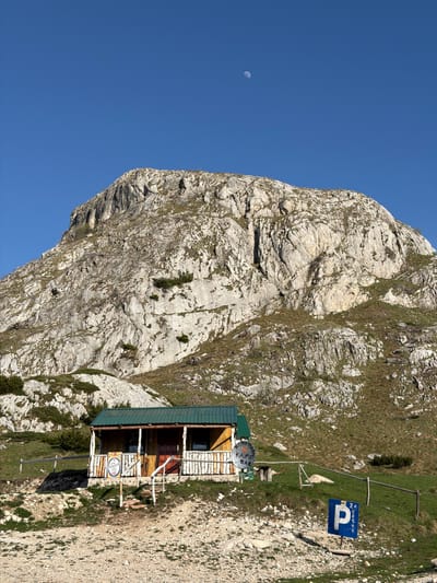 The image depicts a mountain scene in Boričje, Montenegro, featuring a rocky mountain face under a clear blue sky with a visible crescent moon. A wooden structure, likely a mountain hut or cafe, sits on the mountainside, adorned with signs, including a "parking" sign in Cyrillic. The surrounding terrain is a mix of rock and sparse vegetation.