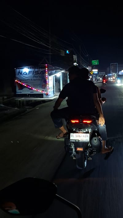 The image captures a nighttime street scene in Blangpeuria, Indonesia. In the foreground, two individuals are riding on a motorcycle, viewed from behind. The driver, wearing a dark shirt and pants, is operating the vehicle, while a passenger sits behind them. The motorcycle's rear taillight is illuminated, and a license plate with the characters "BL 5061 KBK" is visible. The environment includes a street with a slight incline, and on the left side of the road, there is a stall or building with bright, colorful lights and text that appears to read "NAFISA." In the background, other vehicles are present on the road, with their headlights and taillights creating streaks of light. Overhead, a complex network of power lines is visible against the dark night sky. The overall impression is that of a bustling, illuminated street at night in a residential or commercial area.