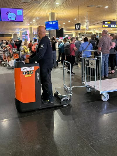 The image captures a scene inside a modern airport terminal, likely Oslo Airport Gardermoen, Norway, given the signage. In the foreground, a bald man wearing a dark blue uniform with an "M" logo on the sleeve is operating an orange and black baggage tug. The tug has a license plate that reads "20017766" and "TCR MAA 1". Attached to the tug is a metal cart with a "Menzies Aviation" logo. The man is looking forward, seemingly focused on his task.

Behind the tug, a crowd of travelers is visible, with many people pulling luggage or standing in queues. The terminal features a wooden ceiling with suspended lighting and displays. A large digital screen on the left displays a cartoonish illustration of a toaster. Another screen further back shows flight information. Signs with numbers and text, including "Ut Exit" (Exit), are visible, indicating navigation within the airport. The floor is a dark, polished surface, reflecting the overhead lights and the surrounding environment. The overall atmosphere suggests a busy travel hub.