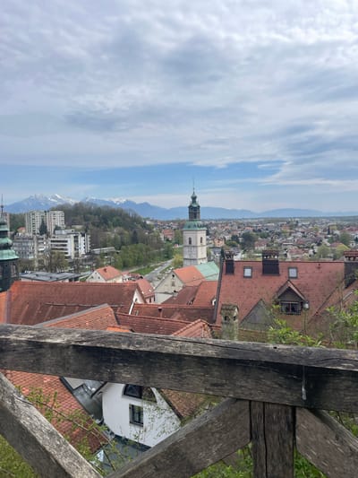 The image shows a view of Skofja Loka, Slovenia, from an elevated vantage point, possibly from the castle. In the foreground, a wooden railing obscures part of the scene. Beyond the railing, the cityscape is visible, characterized by buildings with red-tiled roofs. A prominent church tower with a green spire rises above the surrounding structures. In the distance, mountains are visible against a partly cloudy sky.