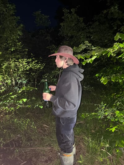 A person wearing a cowboy hat, dark hoodie, dark pants, and rubber boots stands in a grassy, wooded area, possibly in Krasnyi Jar, Russia. The person is holding a green glass bottle. The setting appears to be at night, with limited ambient lighting. Vegetation includes grass, saplings, and mature leafy bushes/trees. The sky in the upper left corner is dark.