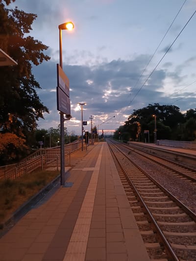 The image depicts a train station platform in Germering, Germany, at dusk. The sky is filled with clouds, with hints of sunset colors peeking through. A single, illuminated street lamp stands on the platform, casting a warm glow on the brick-paved walkway. To the left of the platform, there are trees and a staircase leading down. The right side of the image shows train tracks stretching into the distance, with overhead power lines visible against the sky. A few other street lamps are visible further down the platform. The scene is quiet and appears to be devoid of any people or activity.