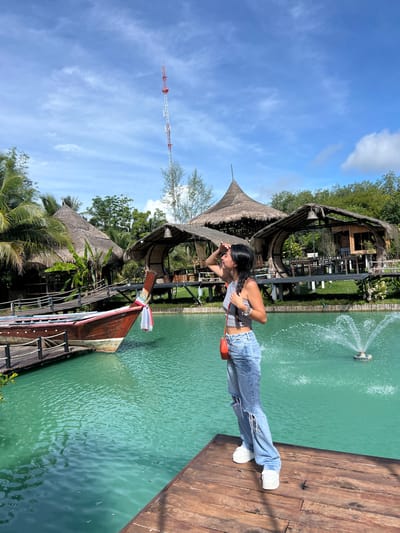 The image depicts an outdoor scene in Tambon Pa Kae Bo Hin, Thailand, featuring a woman standing on a wooden dock. She is wearing ripped light-wash jeans, a grey tank top, and white sneakers, and is holding an orange purse. She is shielding her eyes with her hand as she looks out over a body of turquoise water. In the background, several thatched-roof structures are visible, along with lush green vegetation and palm trees. A wooden boat with a red hull is docked to the left, and a fountain sprays water in the middle of the water. A tall, red and white communication tower stands in the distance against a blue sky with scattered clouds.