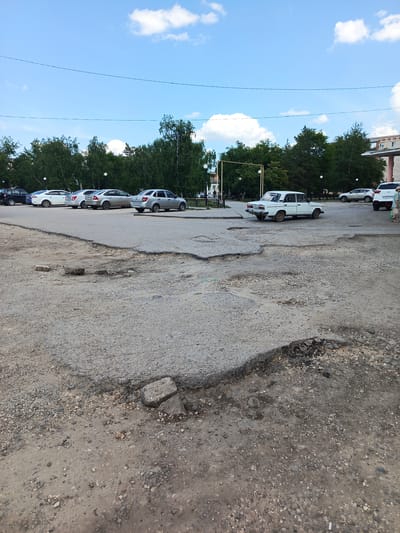 A daytime scene in Uryupinsk, Russia, depicts a damaged asphalt road in the foreground, characterized by numerous potholes and loose gravel. Several cars are parked on a smoother section of road in the background. To the left, a white sedan is parked next to a silver sedan and other vehicles. Further left, a black car is visible. In the middle ground, a white classic sedan is parked, and to its right, a white SUV is parked, with a person standing nearby. Trees form a dense line along the back of the scene, with some buildings visible above the treeline on the right. The sky is blue with scattered white clouds, suggesting fair weather. The overall impression is of a slightly worn urban environment with a focus on the road conditions and parked vehicles. No visible text is discernible.