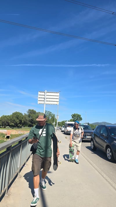 Two men with skateboards are pictured outdoors on a sunny day in Santa Perpètua de Mogoda, Spain. The image is taken from a slightly elevated perspective, looking down and along a paved path next to a road.
In the foreground, a man stands on the paved path beside a metal railing. He is wearing a dark green t-shirt with a white logo, olive-colored cargo shorts, a two-tone baseball cap, white socks, and green-and-white sneakers. He holds a black skateboard under his left arm and is looking down at a black smartphone held in his left hand. A black shoulder bag strap crosses his chest. His prominent shadow falls on the path, indicating strong sunlight.
Further along the path, closer to the road and parked cars, another man is visible. He wears a light-colored t-shirt with a graphic print, light shorts, a blue beanie, white socks, and blue sneakers. He holds a skateboard with a green graphic design under his arm and is leaning against a parked black car. He appears to be looking towards the camera, wearing sunglasses.
The setting includes a road with several parked cars, including a black car whose license plate "C 4973 MC" is partially visible. Between the two men stands a white directional sign with multiple horizontal panels. The background features lush green trees and distant hills or mountains under a bright blue sky, with visible wispy clouds and airplane contrails. The clear sky and strong shadows suggest it is midday or early afternoon. The activity appears to be a casual break during a skateboarding session.