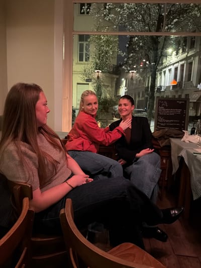 The scene depicts three adult females seated inside a commercial establishment, likely a restaurant or bar, at night in Ixelles - Elsene, Belgium. A large window behind them offers a view of a well-lit city street at night, featuring traditional European buildings, streetlights, a tree with white blossoms, and distant vehicles.

The female on the left, with long brown hair, is seated in a wooden chair, seen in profile looking towards the right. She wears a light-colored, short-sleeved top and dark pants. A gold-colored bracelet and a ring are visible on her right hand.

The central female has blonde hair and wears a red long-sleeved shirt and blue jeans. She is seated, smiling towards the camera. Her right hand is placed on the neck of the female to her right.

The female on the right has dark hair pulled back and wears a dark blazer over a dark top and blue jeans. She is also seated, smiling towards the camera. Her left hand rests on her lap, and she wears black shoes.

Through the window, an outdoor menu board is visible on the right. A table with a striped tablecloth, a dark bottle, and glasses is partially visible beside the menu board. The street scene includes a circular street sign and a rectangular blue street name sign.