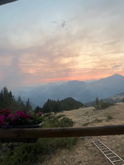 This image captures a panoramic view of a mountain range at sunset, with a foreground featuring a wooden railing, a planter box filled with vibrant pink flowers, and dry, autumnal vegetation. A wooden ladder lies on the ground in the lower right corner. The sky is painted with hues of pink, orange, and pale blue, transitioning into a hazy blue expanse over the layered mountains in the distance. This scene is likely situated in or near San Juan de Plan, a municipality in the province of Huesca, Aragon, Spain, given the characteristic Pyrenean landscape. The environment suggests an outdoor setting, possibly a viewpoint or a balcony overlooking the valley.