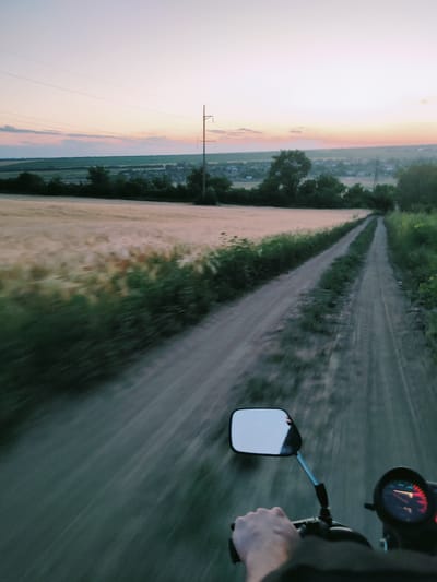 This is a first-person perspective, point-of-view shot of someone riding a motorcycle along a dirt road at dusk. The setting appears to be rural Moldova, possibly near Filipeni.

**Subjects:** The primary subject is the motorcycle's handlebar and the rider's hand gripping it. A rearview mirror is prominent, reflecting a sliver of the sky and possibly the rider or another person. The background features a vast, open landscape with fields of ripe grain, interspersed with trees and low-lying vegetation. In the distance, a small village or settlement is visible, with houses and a few power lines stretching across the horizon.

**Activity/Event:** The image captures the sensation of motion as the motorcycle travels down the dusty track. The blurred effect of the foreground and sides emphasizes the speed and forward momentum.

**Notable Details:** The time of day is clearly dusk, with a soft, warm glow in the sky, transitioning from pale pink to a lighter hue towards the zenith. The weather appears calm and pleasant. The dirt road is well-trodden, with patches of grass and vegetation growing along its edges and in the center. The fields suggest a harvest season. The overall mood is one of peaceful travel and exploration of the countryside.

**Visible Text:** There is no discernible text in the image. The dashboard of the motorcycle has a speedometer with a red needle, but no legible numbers or labels are visible.