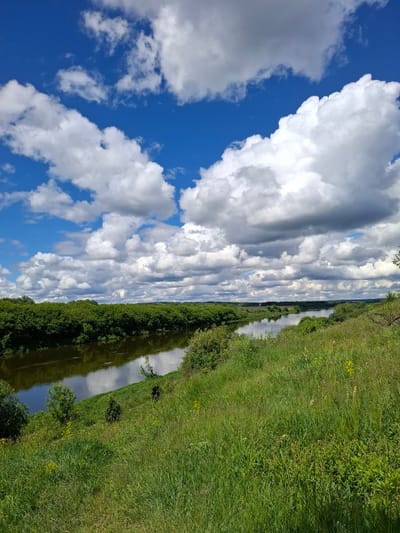 The image captures a rural landscape near Elets-Malanino, Russia. A narrow, winding river or canal reflects the sky, bordered by dense green vegetation on both sides. The sky is mostly blue with large, puffy cumulus clouds, creating a dynamic contrast between light and shadow. The vegetation includes grasses and trees. There are no visible people or animals. The scene is tranquil and depicts a typical rural environment in this region.