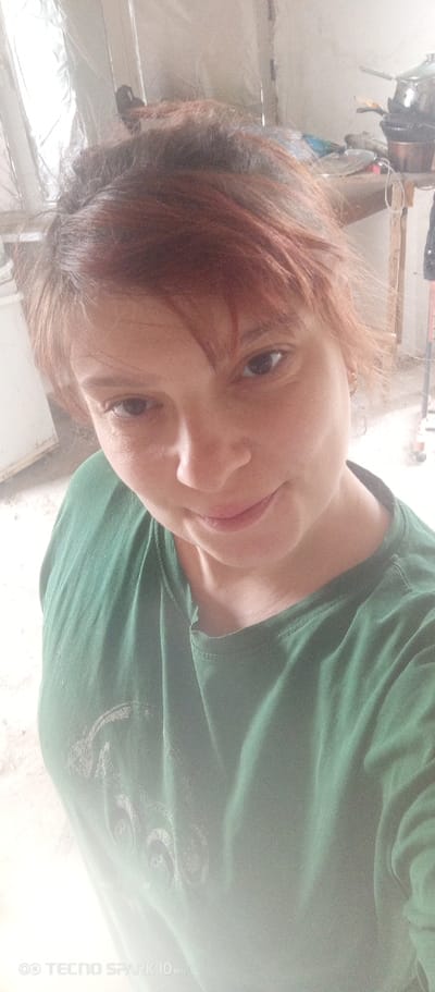 A woman with reddish-brown hair and a green t-shirt is taking a selfie. She is indoors, with a cluttered workspace or storage area visible in the background. The location might be Bereg Kubani, Russia.