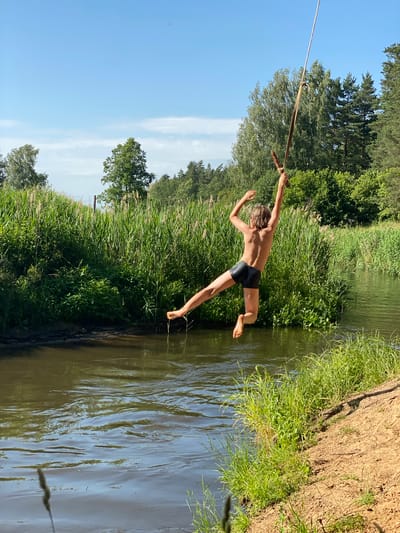 A young boy is captured mid-air as he swings on a rope over a body of water. He is wearing black swim trunks and appears to be in the process of jumping into the water. His body is arched, with his arms extended outwards for balance and his legs positioned as if about to land. The rope he is holding is thick and appears to be made of natural fibers.
The scene is set in a natural outdoor environment. Lush green grass and reeds line the bank of the river, with tall trees forming a dense backdrop. The water is calm and reflects the surrounding greenery and the clear blue sky. A dirt bank with sparse vegetation is visible on the right side of the frame, suggesting a slightly elevated point from which the swing is launched. The sunlight suggests it is a bright, clear day, likely during the afternoon given the angle of the light. The overall atmosphere conveys a sense of fun, adventure, and carefree summer activity. There is no visible text in the image.