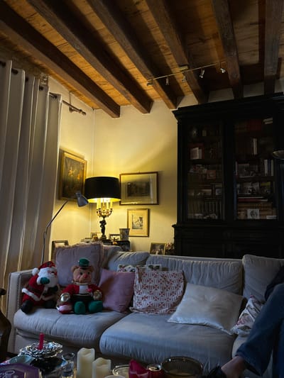 This image captures a warm and cozy indoor scene within a home in Étréchy, France, likely a living room connected to a staircase. The perspective is slightly elevated, looking down into the space.

On the left, a prominent rustic wooden staircase ascends, featuring exposed, heavy timber beams and steps. A modern, multi-spotlight fixture is attached to the wooden structure. The lower section of the staircase appears to be painted white with a slender metal handrail.

The right side of the image reveals a comfortable seating area. A light-colored, upholstered sofa or armchair is visible, on which a person's legs, wearing blue jeans, are resting. The sofa is decorated with several cushions, including one with a red and white geometric pattern, a plain pink one, and a white satin-like cushion. Two festive, plush figures, an elf and a Santa Claus, are seated on the sofa, suggesting a holiday theme.

In the background, a large, dark wooden bookcase with glass doors is filled with books and various decorative objects. The wall behind the sofa is adorned with multiple framed artworks and photographs. The room is illuminated by artificial light, notably from a floor lamp with a black shade casting a warm glow, and what appears to be part of an ornate chandelier. A smaller, metallic task lamp is positioned near the floor. Other decorative items near the sofa include candles, a silver tray, and a small mirror.

The overall impression is one of a relaxed and inviting domestic setting, likely in the evening given the reliance on artificial lighting. No discernible text is visible in the image.