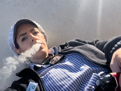 A person with short dark hair is lying on a light gray concrete surface. They are wearing a white and gray striped shirt, a black zip-up jacket, sunglasses, and a white crocheted hat with a dark gray brim. A cloud of smoke is emanating from their mouth. A white card with a green cross emblem is visible on the jacket. The location is outdoors. The city and country of Mensdorf, Luxembourg cannot be determined from this image alone.