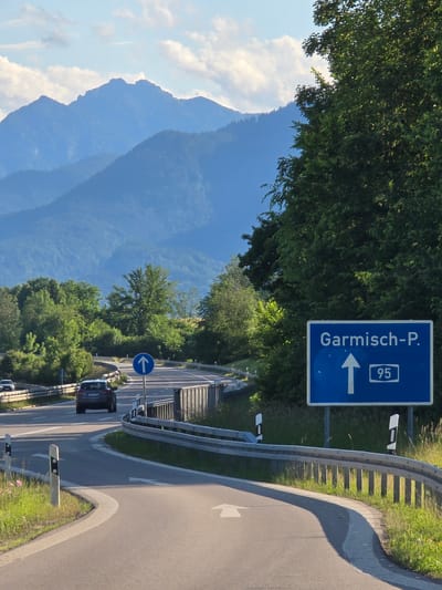 The image presents a detailed view of a road junction in Ohlstadt, Germany, set against a backdrop of distant, layered blue mountains under a partially cloudy blue sky. A multi-lane asphalt road dominates the foreground, curving both to the left and right, with a prominent white arrow painted on the asphalt indicating a straight direction. A dark SUV is visible driving away from the viewer on the main road in the middle ground, with a lighter-colored car further back on an elevated section.

On the right side of the road, a large blue rectangular sign displays "Garmisch-P." with a white upward arrow and the number "95" in a white box, indicating the direction to Garmisch-Partenkirchen via Route 95. A circular blue sign with an upward arrow, signifying a mandatory direction, is positioned between the road lanes. Metal guardrails line the curves of the road and bridges, complemented by reflective black and white guide posts. Green grass and some pink wildflowers are visible along the road shoulders.

To the right, a large, dense green tree occupies a significant portion of the frame, with additional trees and foliage lining the road and obscuring parts of the landscape. The distant mountains, characteristic of the Bavarian Alps, exhibit atmospheric perspective, appearing progressively lighter blue with distance. The scene is illuminated by sunlight, casting long shadows from the guardrails and trees, suggesting either morning or late afternoon. No individuals are depicted in the scene.