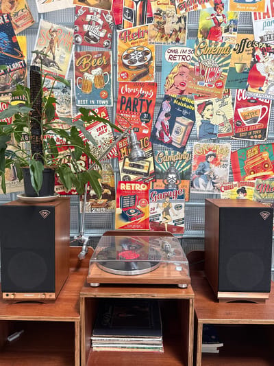 The image depicts a close-up of a collection of vintage-style posters displayed on a wall, with a plant and audio equipment in the foreground. The posters feature various designs, including advertisements for music festivals, beer, cinema, coffee, and travel, all rendered in a retro aesthetic with vibrant colors and illustrative styles. In the foreground, a potted plant with large, green leaves sits on a white saucer. To the right of the plant are two wooden speaker boxes, one on each side of a turntable. The turntable, with a clear dust cover, is placed on a wooden shelf above a stack of vinyl records. The overall scene suggests a curated collection of retro memorabilia and audio equipment. The location is not specified in the image.