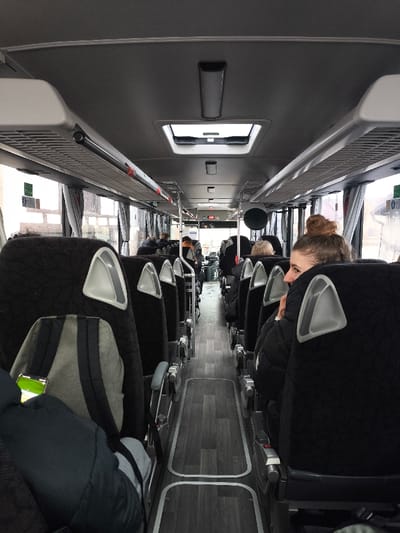 The image displays the interior of a passenger bus in Meistratzheim, France, viewed from a slightly elevated position within the cabin, looking forward down the central aisle. The environment consists of rows of dark-colored, high-backed passenger seats, each featuring light grey triangular accents on the upper backrests. A central aisle, paved with a dark wood-patterned laminate floor bordered by light grey trim, runs the length of the bus. Overhead luggage racks, air vents, and recessed lighting fixtures are visible along the grey ceiling.
Several individuals are present, seated on both sides of the aisle. In the immediate right foreground, a young female with light brown hair styled in a bun is partially visible from the shoulders up, turned to her left, wearing a black quilted jacket. Her face is mostly obscured by the seat in front, but she appears to be looking towards the left side of the bus. On the seat directly to the left in the foreground, a light grey backpack with black straps is positioned, topped with a smartphone displaying a bright green screen. Further down the aisle, other passengers are indistinctly visible, primarily from their backs or partially, occupying seats on both sides. The bus windows on either side show blurred glimpses of an exterior environment, with vague structural elements. No direct interactions between individuals are discernible.
