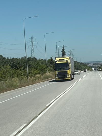 A bright yellow semi-truck with "AUTO EXPERT" written on its side is driving on a paved road. The truck is carrying a grey trailer and is surrounded by other vehicles, including a white car and several motorcycles. The road is bordered by a lush green forest on the left and a clear blue sky with some electrical towers in the distance. The scene appears to be in an outdoor, rural or suburban setting, possibly near the city of Tavri, Greece, given the presence of electrical infrastructure typical of such areas. The lighting suggests it is daytime.