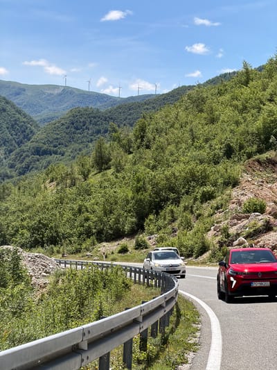 This media file presents a scenic view of a winding mountain road in Šavnik, Montenegro, captured on a clear, sunny day. The image features a two-lane asphalt road curving through a verdant, mountainous landscape.
In the foreground and midground, two cars are visible on the road. A red, modern passenger car is positioned further along the road, with its front end facing away from the viewer. Its license plate, "PG-NN201", is clearly legible. Closer to the viewer, a white passenger vehicle is situated on the right side of the road, adjacent to a metal guardrail. The white car's license plate reads "PG-BR584".
The road is bordered by dense green foliage, with steep, forested slopes rising on both sides and extending into the background. On a distant mountain ridge, several white wind turbines stand tall against the horizon, indicating a wind farm. The sky above is a bright, clear blue, dotted with scattered white clouds, suggesting pleasant weather conditions. The strong sunlight illuminates the scene, highlighting the details of the cars, road, and the rich green vegetation. The perspective offers a view along the road as it traverses the mountainous terrain.