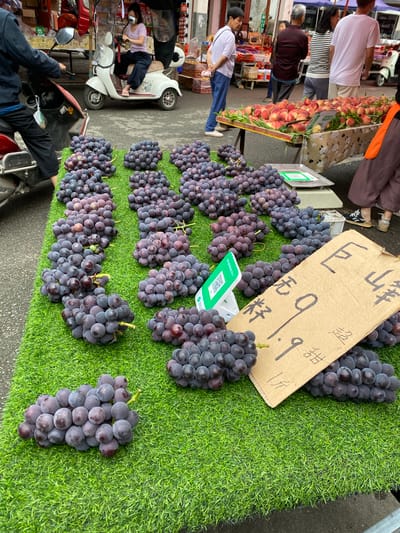 The image captures a bustling street market scene in Chongzhou City, China, likely in an outdoor setting. The foreground is dominated by a vibrant display of dark purple grapes arranged in clusters on a green artificial turf surface, mimicking grass. The grapes appear fresh and ripe, with some bunches showing a purplish-red hue.

To the right of the grapes, a piece of cardboard serves as a price tag. Black handwritten Chinese characters are visible on the cardboard, indicating pricing information. Among these characters, "1.9" is prominently displayed, suggesting a price of 1.9 units of currency per a certain quantity of grapes. Other characters, such as "甘" (sweet) and "斤" (jin, a unit of weight), are also present.

Behind the grapes, a stall with a beige cloth patterned with cartoon sheep is visible, supporting a large quantity of peaches. These peaches are a vibrant blend of red and yellow, suggesting they are also ripe and ready for sale. A silver weighing scale with a green QR code sticker is placed near the grapes, indicating a transaction or payment method.

In the background, various individuals are engaged in market activities. On the left, a person is riding a red and white electric scooter. A woman wearing a light purple top and dark pants is seated on the scooter, her face partially obscured by a white mask. Further in the background, other market-goers can be seen browsing the stalls. The overall environment suggests a lively and active marketplace with fresh produce being sold.