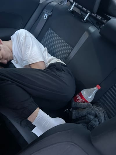 The image shows a person sleeping in the back seat of a car. The person is wearing a white t-shirt and black pants, with white socks. A bottle of Coca-Cola and a gray blanket are on the seat next to the person. The car appears to be parked, and the background suggests an indoor setting, possibly a garage or parking lot. There are no visible indications of the specific city of Krymsk, Russia, in the image.