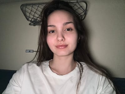 The image depicts a female individual within a train carriage, located in or near Kargat, Russia. The subject, a young adult female, is positioned centrally in a frontal portrait, looking directly into the camera. She has long, dark brown hair, fair skin, dark eyes, and full lips which are slightly parted, exhibiting a subtle sheen. She is wearing a plain white crew-neck t-shirt and a thin, rose-gold colored necklace with a small, discreet pendant. A small, golden earring is visible on her right earlobe. The background consists of a plain, light grey wall. A silver-colored metal mesh luggage rack is mounted on the upper left portion of the wall, and a grey rectangular light fixture is attached to the wall to the left of the subject's head. A dark blue or black surface, likely part of a seat, is partially visible in the lower right portion of the frame. The lighting is even, consistent with an indoor artificial source, illuminating the scene without harsh shadows.