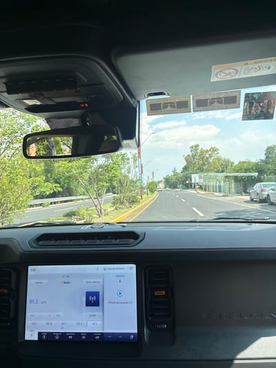 The image shows the interior of a car, likely a Ford Bronco, driving along a road in Tepeji de Ocampo, Mexico. The dashboard is visible, with a central display showing a radio station tuned to 91.3 FM. The rearview mirror reflects a view of the road and surrounding greenery. An airbag warning label and stickers are visible on the sun visor. The display screen shows a menu in Spanish.