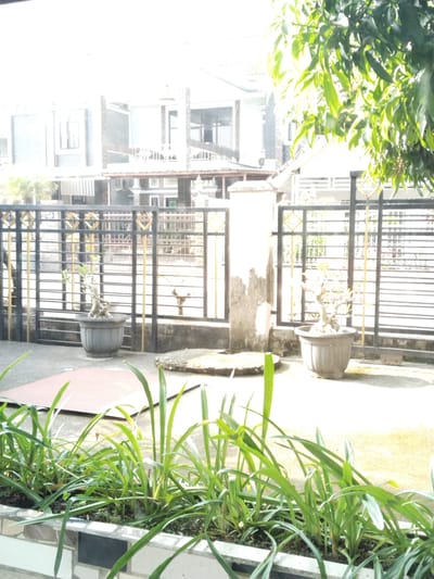 The image depicts a residential area with a fence in the foreground, a house in the background, and trees. In the front, there is a planter box filled with green plants. Behind the planter, a black metal fence with gold diamond accents runs horizontally across the frame. Two potted bonsai trees are visible behind the fence. A tall, cylindrical concrete pillar stands to the left of center, weathered and stained. The house in the background appears to be a modern structure with multiple stories, featuring windows and balconies. Lush green foliage from trees overhangs the scene from the upper right, suggesting a tropical environment. The lighting indicates it is daytime, with strong sunlight creating bright highlights and some shadows. The scene is likely located in a residential neighborhood within the city of Lahat, Indonesia, characterized by its warm climate and abundant vegetation. No people or specific actions are discernible in the image.