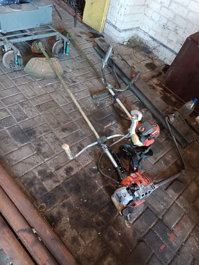 The image depicts a cluttered storage area with multiple lawn maintenance tools and materials. In the foreground, two strimmers and a leaf blower are laid out on a tiled floor. One strimmer, orange and grey, is partially disassembled, with its engine and shaft separated. The other, longer strimmer, is intact and lies alongside it. A red and grey leaf blower is positioned in the lower right of the frame. In the background, a wheeled cart with a large, round cutting head is visible, along with various pipes and a metal shelf. The environment appears to be an indoor space, possibly a garage or workshop, with brick walls and a concrete floor. The overall impression is one of disarray and disuse of the equipment. This scene is located in Vuhlehirsk, Ukraine.