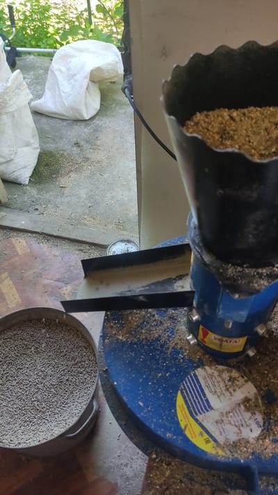 This image depicts a close-up of a grain grinder or pelletizer, likely used in an agricultural or animal feed context. The machine is primarily blue and appears to be made of metal. A wide, funnel-like chute at the top is filled with a light brown granular material, presumably grain or feed. The chute leads into the main body of the machine, which has visible bolts and a yellow warning label. A circular sticker with text and graphics is affixed to the blue casing. The machine is positioned outdoors or in an open-air setting, as suggested by the concrete ground scattered with dust and debris, and glimpses of greenery in the background. To the left, two white, woven sacks are visible, one partially filled and the other appearing empty. In the foreground, a round metal container is filled with a large quantity of small, pelletized feed. The overall impression is one of an industrial or farm-related workspace, possibly during daylight. The text on the circular sticker includes the words "2-100" and what appears to be "EAC" and some Cyrillic script, suggesting a product model number and certification mark. The context of Mohyliv-Podilskyi, Ukraine, further supports the agricultural interpretation. No people are visible in the frame.