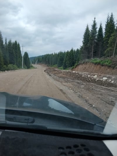 The image depicts a dirt road winding through a forested area with scattered telephone poles. The road appears to be under repair or has been affected by a landslide, with debris and rocks present along the side. The terrain is uneven, and the surrounding vegetation consists of dense evergreen trees. The sky is overcast, suggesting a cloudy or possibly rainy day. The foreground shows a portion of a vehicle's dashboard and windshield, indicating the photo was taken from inside a car. The location is identified as a road in the vicinity of Toktogul, Kyrgyzstan.