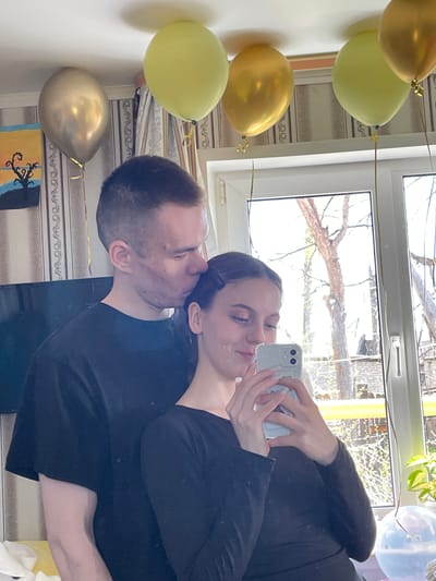A couple is taking a selfie indoors, with a window visible behind them. A string of gold and light green balloons is attached to the wall on the left. Outside the window, trees and buildings are visible. The location is likely Ulukulevo, Russia, as per the prompt instructions, although this cannot be verified from the image itself.