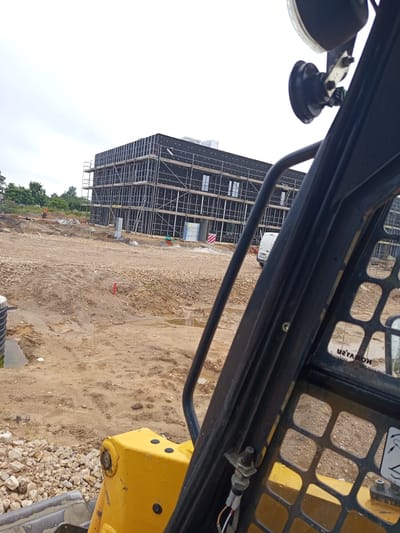 The image depicts a construction site in Preiļi, Latvia. In the background, a multi-story building is under construction, covered in black insulation material and surrounded by scaffolding. The foreground is dominated by a yellow construction vehicle, possibly a skid steer loader, partially obscuring the view. The ground is uneven and consists of dirt, gravel, and puddles of water. Several construction elements, such as pipes and barriers, are visible. The sky is overcast.