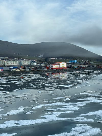 The image depicts a view of the city of Pevek, Russia, as seen from the water. In the foreground, the dark water is dotted with numerous ice floes of varying sizes and shapes. The ice is a mixture of white and grey, with some floes appearing thicker and more solid, while others are thinner and more translucent.
Behind the water, the city of Pevek is visible. The cityscape features a cluster of multi-story buildings, many of which are painted in bright, contrasting colors such as red, yellow, white, and blue. These buildings appear to be residential or administrative structures. Scattered among them are smaller, industrial-looking buildings, warehouses, and shipping containers. One notable building has a red, domed roof, suggesting a church or other significant architectural feature. A prominent building in the mid-ground is a bright orange and white structure, possibly a warehouse or industrial facility. Further back, the land rises into a large, dark, and rounded mountain, partially shrouded in clouds. The overall impression is of a coastal settlement in a cold, possibly Arctic, region, with a mix of colorful urban development and a harsh, natural environment. The sky is overcast and a pale blue, with heavy clouds obscuring the upper portions of the mountain.