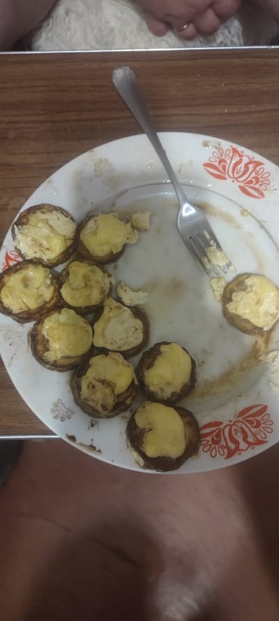 The image shows a plate of food on a wooden table. The plate is white with an orange floral pattern. On the plate, there are several circular pieces of food, possibly mushrooms, topped with melted cheese. A fork is also present on the plate. In the background, a person's hand with a ring is visible, resting on a fluffy white surface. The environment is likely a domestic setting, but there is no specific information to place it in Svitlovodsk, Ukraine.