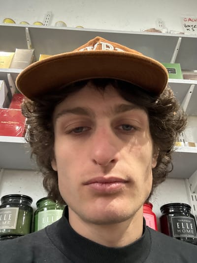 A close-up, front-facing photograph depicts a young adult Caucasian male, approximately 20-30 years of age, with dark curly hair and blue eyes, wearing a dark-colored crewneck shirt and a brown baseball cap with white lettering worn backward. His facial expression is neutral. The subject is situated indoors within what appears to be a retail or storage environment in Joinville-le-Pont, France. Behind the subject, white shelving units are stocked with various commercial products. On the left upper shelves, multiple packaged boxes are visible, including items labeled "MAUBOUSSIN" and "PASCAL MONACO JARDIN RED." On the right upper shelf, a handwritten label on a piece of paper reads "CALN BOXER K3." On the lower shelves, behind the subject's shoulders, several glass jars are present: two green-tinted jars are visible to the left, one clearly marked "ELLE HOME Coconut Candle," and a dark-colored jar to the right is labeled "ELLE HOME Vanilla Intense." The scene is illuminated by uniform artificial lighting.