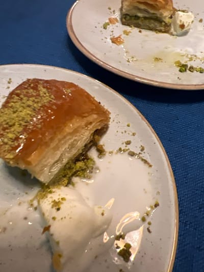 The image depicts a close-up view of two light-colored ceramic plates resting on a dark blue, textured fabric surface. The scene is located in Bağcılar, Turkey.
In the foreground, a single, approximately 8x6 cm piece of baklava is centered on a plate, featuring golden-brown flaky layers, a glossy syrup coating, and a generous topping of green crushed pistachios. Adjacent to the baklava, a melting quantity of white cream or ice cream is visible, intermingled with pistachio fragments and syrup residue. The plate itself shows scattered green pistachio remnants and clear liquid residue.
In the upper right background, a second, partially visible plate holds a smaller, partially consumed piece of baklava, also exhibiting a golden-brown appearance, syrup, and green crushed pistachios. This plate also contains a small amount of melting white cream or ice cream and scattered pistachio fragments.
No individuals or direct actions are visible; the scene presents the aftermath or an paused state of dessert consumption.