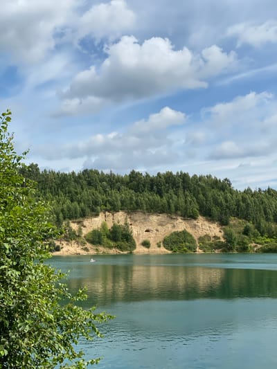 The image shows a serene lake with a sandy cliff face and a dense forest on the opposite bank. In the foreground, the vibrant green foliage of a tree partially obscures the view. The lake's surface is calm, reflecting the cloudy blue sky and the surrounding landscape. A small white raft is visible in the distance on the water, and a few people can be seen on the sandy bank near the cliff. The cliff itself is characterized by its light-colored sand and some patches of greenery. The forest that crowns the cliff is a lush expanse of evergreen trees. The overall impression is one of a natural, tranquil setting, possibly a popular recreational spot.