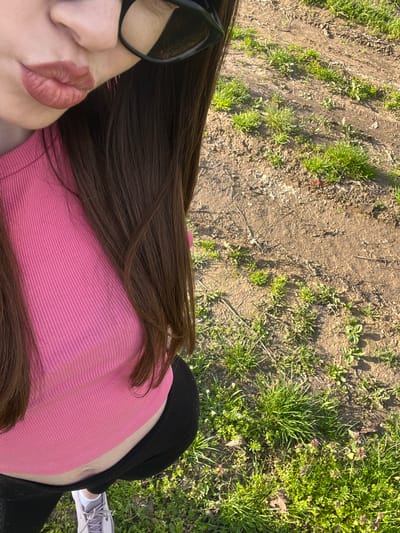A first-person, aerial view image showing a person with brown hair, wearing glasses, a pink shirt, and black pants, sitting on grass and dirt. The location cannot be confirmed as Blaskovo, Bulgaria. A single foot and shoe are visible.